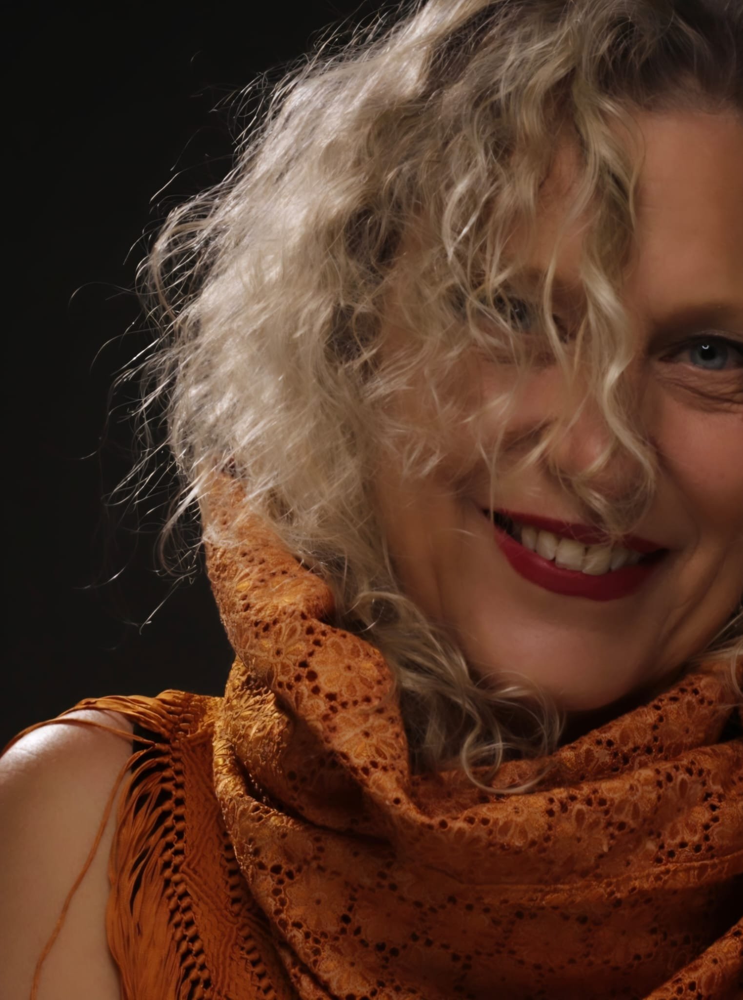 Retrato en color de Mónica Ibáñez Fernández de Lis: melena rizada rubia, sonriendo con los labios pintados de rojo y un pañuelo de encaje naranja, sobre fondo oscuro